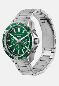 Zelena analogna ura s srebrnim pasom iz nerjavečega jekla; vključuje zeleno teksturirano številčnico, svetleče oznake za ure in prikaz datuma.