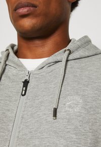 Gray zip-up hoodie with a drawstring hood, featuring a subtle logo on the chest, made of soft, textured fabric. Zipper and metal accents present.