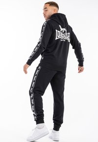 Sudadera con capucha negra y pantalones deportivos a juego, hechos de algodón, que presentan el logo "Lonsdale" y franjas laterales blancas. Estilizado con zapatillas blancas.