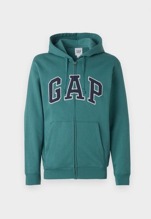 Blågrøn lynlåshættejakke med forlommer, snøretræk i hætten og store marineblå "GAP"-bogstaver med hvid kant hen over brystet.