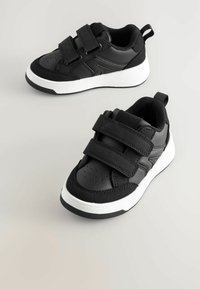 Baskets noires avec deux bandes Velcro, un dessus texturé et une semelle en caoutchouc blanc. Comprend des perforations pour la respirabilité et une languette.