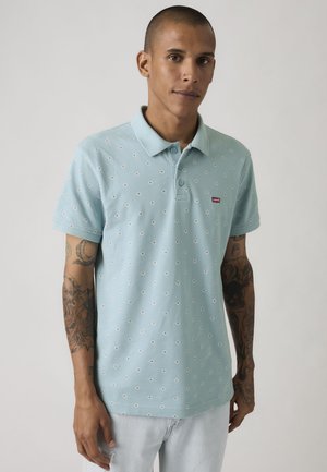 Mężczyzna z tatuażami, ubrany w jasnoniebieską, wzorzystą koszulkę polo oraz jasne dżinsy, stoi na jednolitym tle.