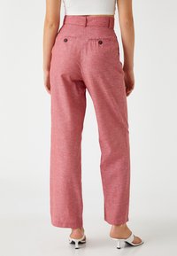 Pantalones rosa texturizados de cintura alta, con piernas rectas y dos bolsillos traseros; combinados con sandalias de tacón blancas.