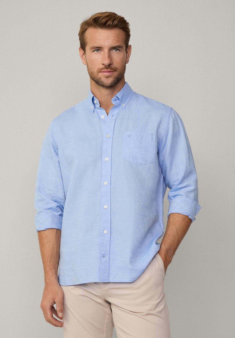 Camisa de botones azul claro hecha de lino, con un bolsillo en el pecho y mangas enrollables. Diseño sencillo con botones blancos y una textura suave.