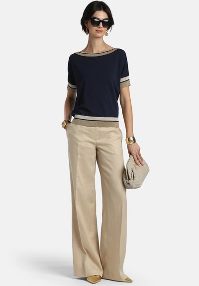 Frau trägt ein marineblaues Oberteil mit beigem Rand, weite beigefarbene Hose, goldene Armbänder, hält eine beige Clutch und trägt schwarze Sonnenbrille.