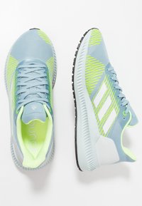 Scarpe sportive azzurro chiaro con tomaia in rete, dettagli verdi, strisce bianche e suola flessibile in gomma nera. Presentano un collare e una linguetta imbottiti.