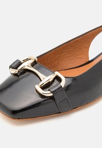 Chaussure plate en cuir verni noir avec un design à bout carré. Présente un grand accent de boucle dorée et une doublure intérieure en cuir beige.