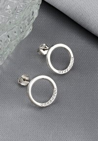 Silverfärgade hoopörhängen med en glänsande yta, prydda med klara kristaller längs ena kanten, placerade mot en texturerad grå bakgrund.