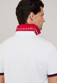 Camisa polo blanca con cuello rojo que presenta el texto "ACONNABLE" y ribetes marinos en las mangas. La tela parece lisa y texturizada.