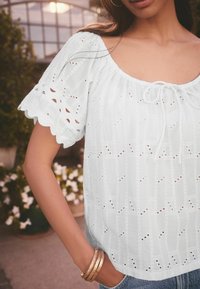Blouse blanche en broderie anglaise avec un décolleté carré, des manches froncées et un détail à nouer, présentant un motif texturé et un design ajouré.