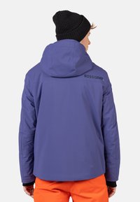 Fioletowa kurtka ocieplana z kapturem, z czarnym logo na ramieniu, noszona z jasnopomarańczowymi spodniami, widoczna tekstura materiału.