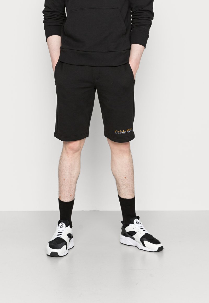 Shorts neri in cotone con tasche laterali, cintura elasticizzata e logo ricamato "Calvin Klein" arancione. Abbinati a sneakers nere e bianche.