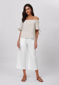 Schulterfreies gestreiftes Oberteil mit gebundenen Ärmeln, kombiniert mit weiter geschnittenen weißen Hosen. Sandalen vervollständigen das Outfit. Neutrale Farben und glatte Texturen.