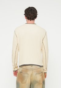 Μπεζ μακρυμάνικο πουκάμισο με υφή, στρογγυλή λαιμόκοψη και αντίθετη καφέ διακόσμηση στο γιακά και στις μανσέτες. Φοριέται με ξεθωριασμένο μπεζ παντελόνι.