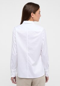 Weißes Langarmhemd mit einem kragenlosen Ausschnitt, glatter Textur und knöpfbaren Manschetten. Der Rücken weist ein sauberes, maßgeschneidertes Design auf.