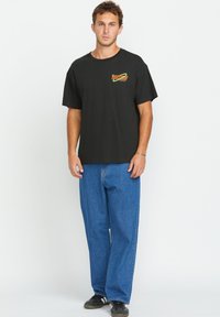 Schwarzes Baumwoll-T-Shirt mit kurzen Ärmeln, mit einem bunten Grafikdesign auf der Brust. Kombiniert mit lässigen blauen Jeans und schwarzen Sneakers.