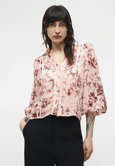 Femme portant un chemisier rose à motifs floraux avec des manches bouffantes et un pantalon noir, posant devant un fond blanc uni.