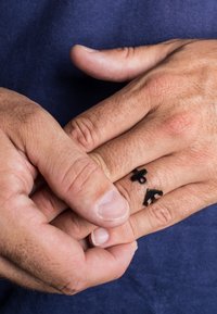 L'anello nero presenta tre forme simboliche: un cerchio, un segno più e una forma triangolare, posizionate su una mano con un aspetto di pelle texturizzata.