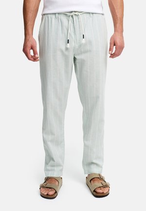 Mann trägt hellgraue Leinenhosen mit Tunnelzug und dezenten Streifen sowie beige Sandalen mit doppeltem Riemen, steht vor weißem Hintergrund.