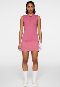 Rosa ärmlös polo klänning med krage och en åtsittande design. Har en liten logotyp på bröstet och en enfärgad textur. Vita sportstrumpor och skor.