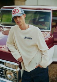 Jeune homme portant une chemise beige à manches longues et une casquette, appuyé sur l'avant d'un pick-up vintage rouge et blanc.