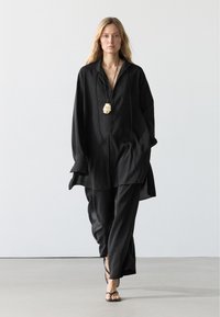 Frau geht, trägt ein locker sitzendes schwarzes Hemd und schwarze Hose, schwarze Sandalen und eine große cremefarbene Anhänger-Halskette an einer schwarzen Schnur.