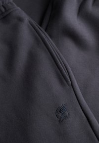 Pantalon en tissu gris foncé avec une poche sur la couture latérale et un petit logo brodé près de la poche.