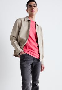 Chaqueta beige con cremallera sobre una camiseta de color coral, combinada con jeans gris oscuro. Diseño sencillo con cuello y puños abotonados.