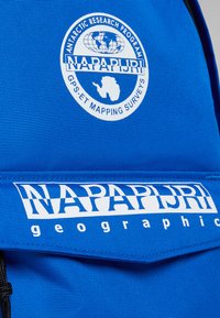 Mochila azul con una tela texturizada. Presenta un logo circular blanco y letras blancas que dicen "NAPAPIRJI geographic" en la parte frontal.