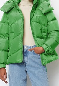 Bright green puffer jacket with a shiny finish, featuring a hood, zip closure, and padded design. Worn over a beige sweater and jeans.