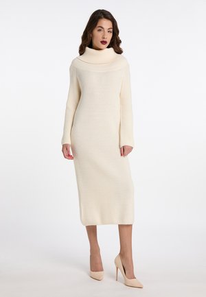 Kvinde iført en cremefarvet ribstrikket midi kjole med bred rullekrave og lange ærmer, kombineret med beige højhælede sko.