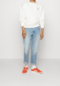 Felpa oversize di colore chiaro con un piccolo logo, jeans in denim blu con risvolti, e sneakers in suede arancione con dettagli bianchi.