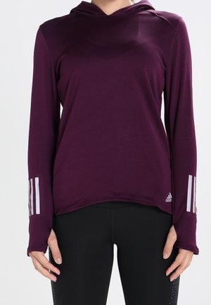 Vrouw die een paarse sporthoodie met lange mouwen en duimgaten draagt, samen met zwarte leggings, staand tegen een effen achtergrond.