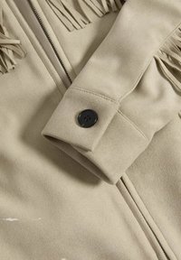 Béžová umělá semišová bunda s předním zipem, třásněmi a černým knoflíkovým detailem na manžetě. Měkká textura s hladkým povrchem.