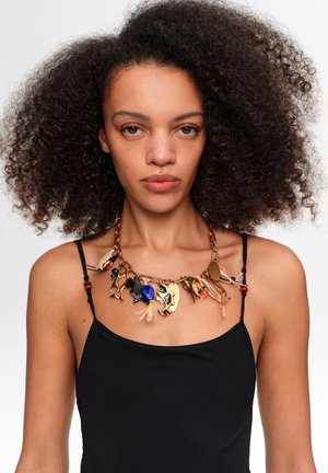 Mujer con cabello rizado que lleva una camiseta negra sin mangas y un collar grueso de cadena dorada con amuletos abstractos y coloridos.