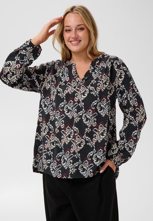 Femme souriante aux cheveux blonds portant un chemisier noir à motifs crème et rouge, posant avec une main touchant ses cheveux et l'autre dans la poche.