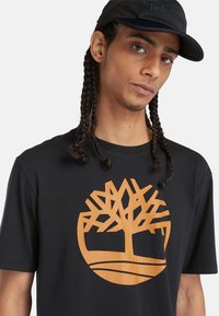 Schwarzes T-Shirt mit kurzen Ärmeln und einem orangen Baum-Logo. Glattes Material, runder Ausschnitt und lockere Passform.