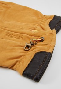 Tan suede handsker med sorte læderdetaljer og en metal spænde med rød og gul snor. Timberland-logo prægede på overfladen.