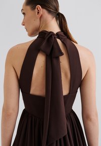 Brown halter dress with a tied bow at the neck, open back, and pleated skirt. Made from soft, textured fabric.
