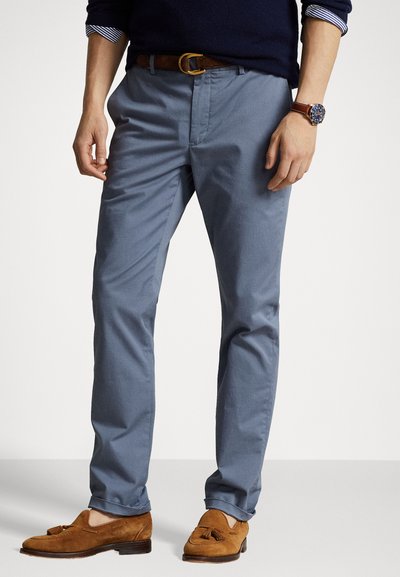 Chino blu deschis din bumbac, cu o croială regulară, buzunare laterale și o curea din piele maro. Mocasinii din suede maro completează încălțămintea.