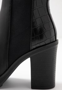 Bottines à talons noirs présentant un mélange de cuir lisse et de matériau texturé en croco gaufré ; panneau latéral élastique ; talon large.