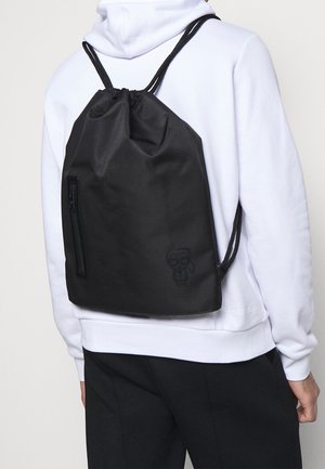 Sac à dos noir avec cordon de serrage, doté d'une texture lisse, comprenant une poche zippée sur le côté et un détail de personnage de dessin animé en relief.