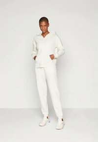 Žena stojí oblečená v bielej mikine s kapucňou, zodpovedajúcich bielych teplákových nohaviciach a bielych teniskách na pozadí jednofarebného svetlého pozadia.