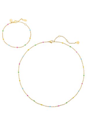 Frau trägt weißen Blazer mit zartem goldfarbenem Kettenhalsband und passendem Armband, verziert mit kleinen bunten Perlen.