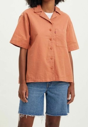 Persona con camisa de manga corta color óxido abotonada y pantalones cortos vaqueros azules desgastados, de pie contra un fondo liso.
