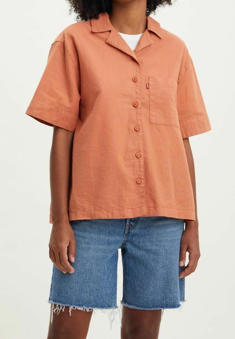Personne portant une chemise à manches courtes boutonnée de couleur rouille et un short en denim bleu effiloché, debout devant un fond uni.