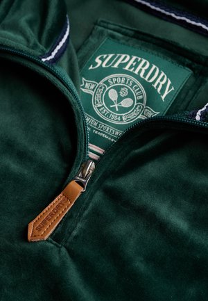 Groene zip-up hoodie met een zachte, fluwelige textuur, voorzien van een leren trekflap, een logopatch en een gestreept kraagdetail.