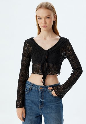 Schwarzes Spitzen-Crop-Top mit langen Ärmeln, V-Ausschnitt und einer vorderen Bindeschleife. Kombiniert mit hochgeschnittenen blauen Jeans. Strukturiertes Blumenmuster.