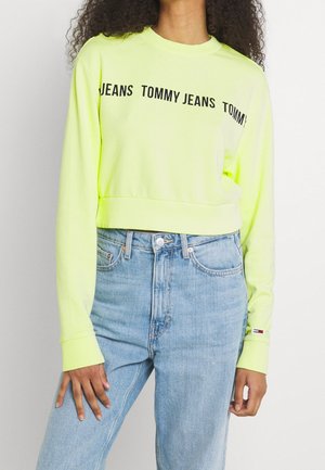 Kvinde iført limegrøn cropped sweatshirt med teksten "TOMMY JEANS" og lyseblå højttaljede jeans, stående mod en ensfarvet baggrund.