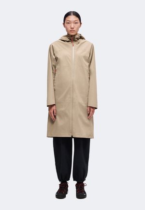 Kvinde iført en beige lang regnfrakke med hætte, sorte bukser med elastiske manchetter og sorte vandresko med røde snørebånd, stående mod en ensfarvet baggrund.